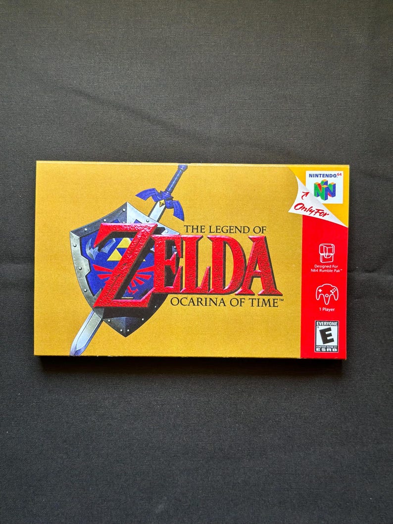 May include: A rectangular gold-coloured video game box for The Legend of Zelda: Ocarina of Time for Nintendo 64. The cover features a blue and silver shield with a sword, the red title text, and a red side panel with the Nintendo 64 logo and ESRB rating.