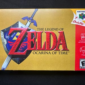 May include: A gold-coloured video game box for The Legend of Zelda: Ocarina of Time for Nintendo 64. The front features a blue and silver shield with a sword, the game title in red text, the Nintendo 64 logo, and an ESRB Everyone rating icon.