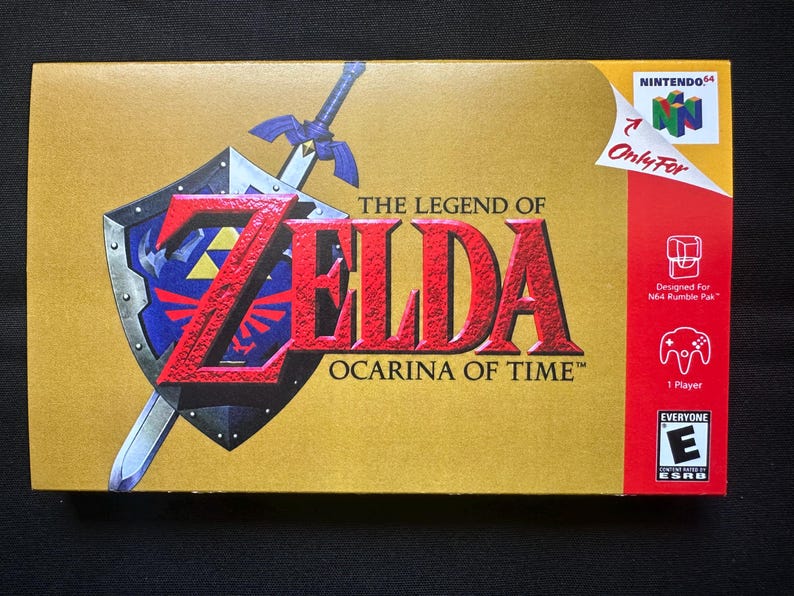 May include: A gold-coloured video game box for The Legend of Zelda: Ocarina of Time for Nintendo 64. The front features a blue and silver shield with a sword, the game title in red text, the Nintendo 64 logo, and an ESRB Everyone rating icon.