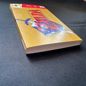 May include: A rectangular video game box for The Legend of Zelda: Ocarina of Time for the Nintendo 64. The gold-coloured cover features a graphic of a sword and shield, with the game title printed in red and black text across the front surface.