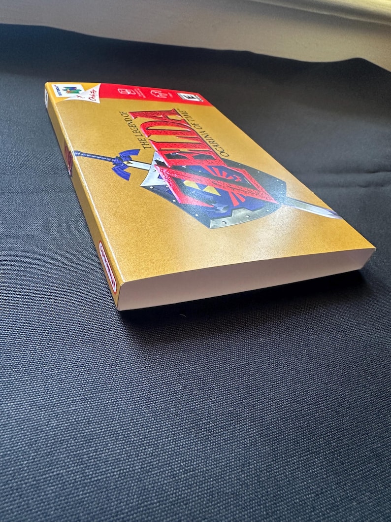 May include: A rectangular video game box for The Legend of Zelda: Ocarina of Time for the Nintendo 64. The gold-coloured cover features a graphic of a sword and shield, with the game title printed in red and black text across the front surface.