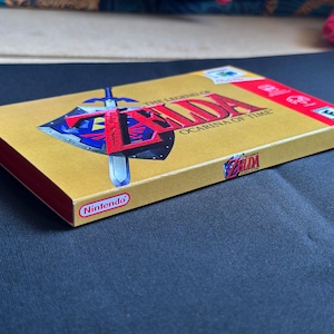 May include: A gold-coloured Nintendo 64 video game box for The Legend of Zelda: Ocarina of Time. The front features the game title in red text, a blue and silver shield with a sword, and an ESRB rating. The Nintendo logo is printed on the side.