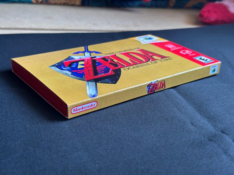 May include: A gold-coloured Nintendo 64 video game box for The Legend of Zelda: Ocarina of Time. The front features the game title in red text, a blue and silver shield with a sword, and an ESRB rating. The Nintendo logo is printed on the side.