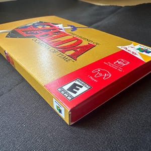 May include: A gold-coloured Nintendo 64 video game box for The Legend of Zelda: Ocarina of Time. The box features a red side panel with an ESRB Everyone rating, a 1-player icon, and a Rumble Pak compatibility graphic, along with the N64 logo.
