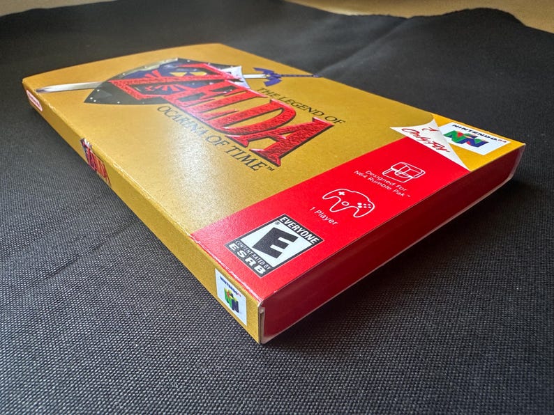 May include: A gold-coloured Nintendo 64 video game box for The Legend of Zelda: Ocarina of Time. The box features a red side panel with an ESRB Everyone rating, a 1-player icon, and a Rumble Pak compatibility graphic, along with the N64 logo.