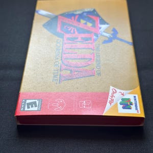 May include: A rectangular gold-coloured video game box for The Legend of Zelda: Ocarina of Time for the Nintendo 64. The front features a shield and sword graphic, while the red bottom section displays the ESRB E rating and console logos.