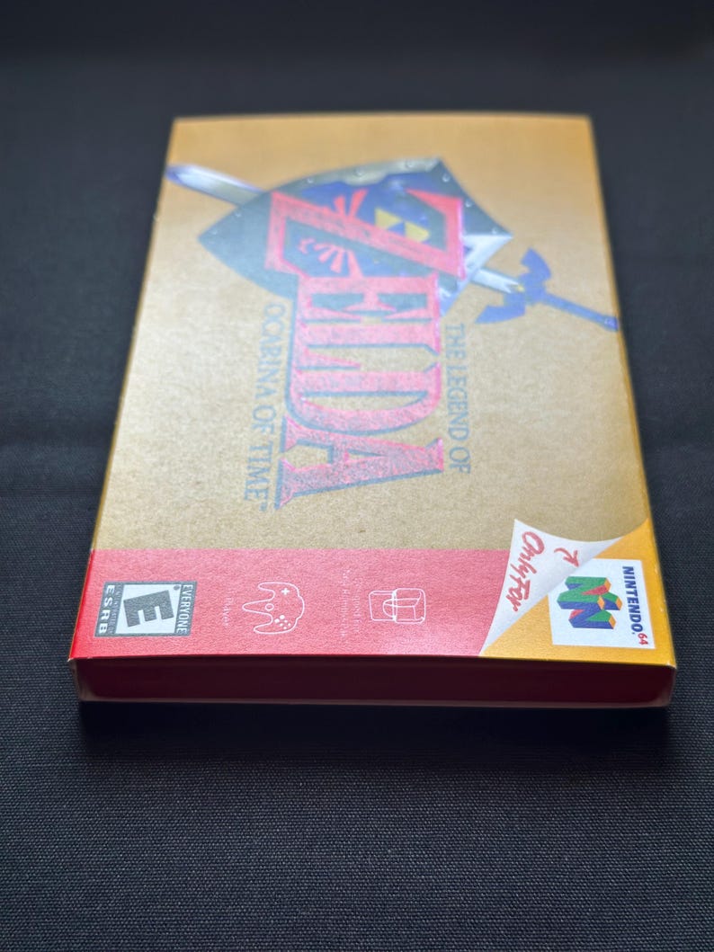 May include: A rectangular gold-coloured video game box for The Legend of Zelda: Ocarina of Time for the Nintendo 64. The front features a shield and sword graphic, while the red bottom section displays the ESRB E rating and console logos.