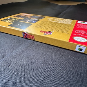 May include: A rectangular cardboard video game box for The Legend of Zelda Ocarina of Time for Nintendo 64. The box is gold with red accents, featuring the game logo, screenshots, and descriptive text on the back. It includes the Nintendo logo.