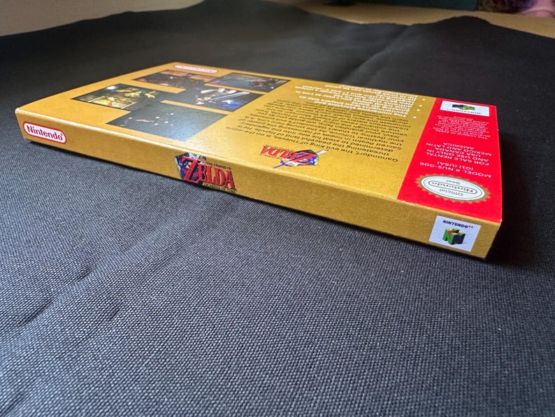 May include: A rectangular cardboard video game box for The Legend of Zelda Ocarina of Time for Nintendo 64. The box is gold with red accents, featuring the game logo, screenshots, and descriptive text on the back. It includes the Nintendo logo.