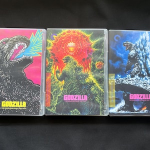 May include: Three Godzilla Blu-ray cases from the Criterion Collection. The left case features a black Godzilla on a pink background. The center case shows Godzilla against a red explosion. The right case depicts Godzilla with blue spikes.