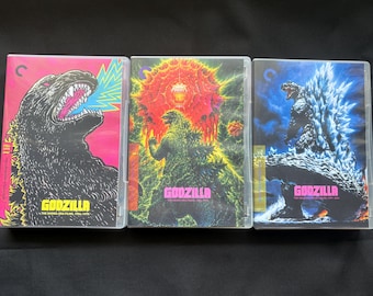 May include: Three Godzilla Blu-ray cases from the Criterion Collection. The left case features a black Godzilla on a pink background. The centre case shows Godzilla against a red explosion. The right case depicts Godzilla with blue spikes.