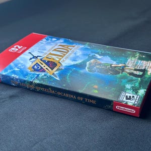 May include: A Nintendo Switch game case for The Legend of Zelda: Ocarina of Time. The cover features a green forest scene with a character, the game title in gold lettering, and the Nintendo Switch logo. The spine displays the game title clearly.