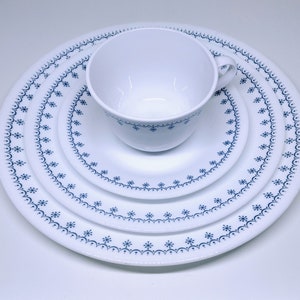 May include: A white dinnerware set with a blue snowflake pattern. The set includes a cup, a saucer, a salad plate, and a dinner plate. The plates are round and vary in size. The cup has a handle.