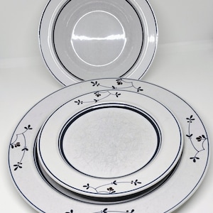 May include: A stack of white ceramic plates with a navy blue trim. The largest plate has a floral design with dark red accents. The plates are arranged on a white background.