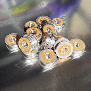 May include: A group of silver and orange bearings. The bearings are arranged in a pile on a silver surface.