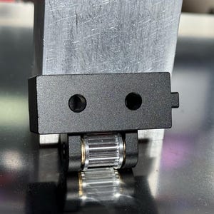Peut inclure: Pièce d'imprimante 3D en métal noir avec un engrenage et deux trous. La pièce est montée sur une surface argentée.