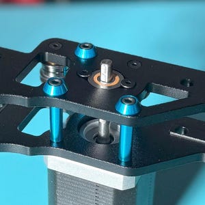 Könnte beinhalten: Eine schwarze mechanische Komponente aus Metall mit blauen Akzenten. Die Komponente hat zwei geschichtete Platten, die durch blaue zylindrische Stützen und blaue Schrauben verbunden sind. Das Objekt befindet sich vor einem hellblauen Hintergrund.