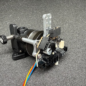 May include: A close-up of a black and silver 3D printer extruder with a silver metal arm and a black motor. The extruder has a black and white wire coming out of it.