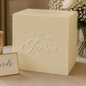 May include: A cream-colored square card box featuring a slot on top and white raised lettering that reads "MR. & MRS. Jansen." Next to the box is a small gold-framed sign with the word "cards" written in black script and two glass candle holders.