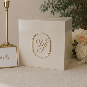 May include: A square, off-white card box with a slot on top and a gold oval emblem featuring the letters TJ on the front. Beside it sits a gold-framed sign with the word cards in cursive script, two tall gold candlesticks, and white flowers.