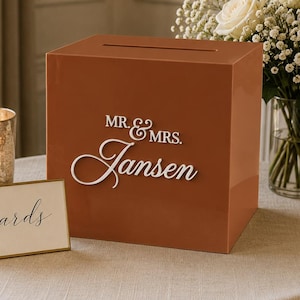 May include: A burnt orange square acrylic card box featuring a horizontal slot on top and white raised lettering that reads MR. & MRS. Jansen. Next to the box is a small rectangular sign with a gold frame displaying the word cards in script.