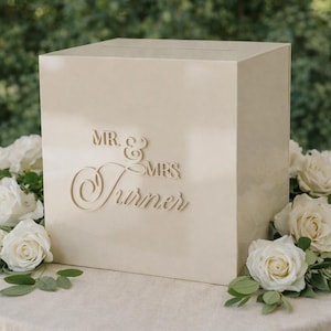 May include: A cream-colored wedding card box with the words "Mr. & Mrs. Turner" engraved on the front. The box is surrounded by white roses and greenery, set on a round table.