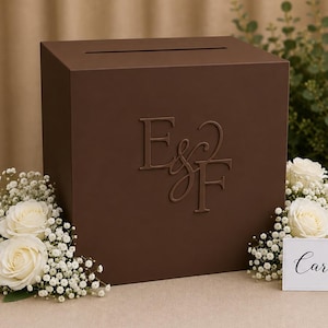 May include: A square dark brown wooden card box featuring a slot on top and the embossed letters E&F on the front. The box is decorated with white roses and baby's breath, accompanied by a small white sign with the word Cards in black script.
