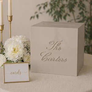 May include: A beige square card box featuring the raised script text The Carters on the front and a horizontal slot on the top. Beside the box is a small rectangular sign with a gold frame displaying the word cards in gold script lettering.