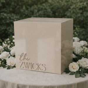 May include: A beige rectangular acrylic card box with a slot on the top surface. The front of the box features the raised white text "the ZWICKS" in a combination of script and capital lettering, positioned on the lower left side of the box.