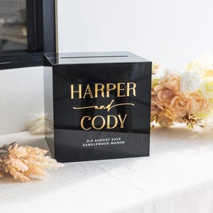 May include: A black acrylic square box with a horizontal slot on top. The front features gold lettering that reads HARPER and CODY, with the text 3rd AUGUST 2025 SANDLEWOOD MANOR printed in white below the names in a smaller font size.