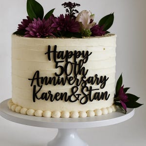 May include: A white frosted cake with the words "Happy 50th Anniversary Karen & Stan" in black script. The cake is decorated with dark purple flowers and greenery, and sits on a white cake stand.