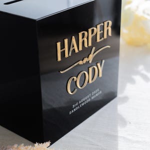 May include: A black rectangular box with a slot on top, featuring raised gold lettering that reads HARPER and CODY. Below the names, smaller white text reads 3rd AUGUST 2025 SANDLEWOOD MANOR. The box has a glossy finish and a clean, modern design.