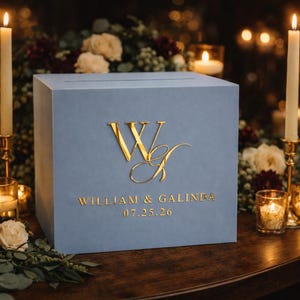 May include: A light blue wedding card box with gold lettering. The box features the initials "W & G" and the names "William & Galinda" with the date "07.25.26". Lit candles and floral arrangements surround the box.