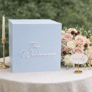 May include: A pale blue card box with the name "The Williams" in white script. The box is on a white tablecloth, beside a floral bouquet and two gold candle holders with lit candles. A small sign reads "cards".