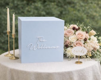 May include: A light blue square card box featuring the white script text The Williams on the front. The box is accompanied by two tall cream-colored taper candles in gold holders and a small oval sign with the word cards in gold script.