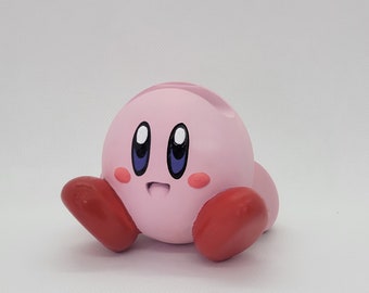 May include: A pink and red character figurine with large eyes and a friendly expression. The character has a round body and short, stubby arms and legs.
