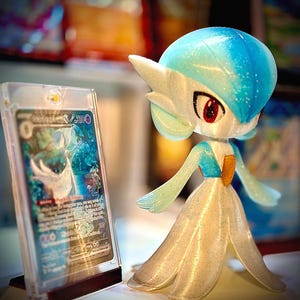 May include: A Gardevoir figurine with a blue and white color scheme, standing next to a trading card in a protective case. The figurine has a flowing dress and detailed features. The card displays a colorful illustration.