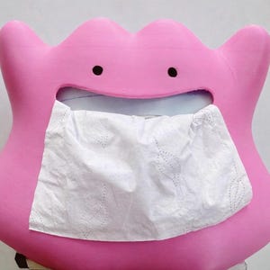 Puede incluir: Un dispensador de pañuelos de color rosa brillante con forma de personaje de dibujos animados con una boca ancha y abierta. El personaje tiene dos pequeños ojos negros circulares y una forma suave y redondeada. Un pañuelo blanco sale por la boca.