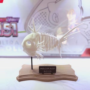 May include: A white Magikarp skeleton figure on a brown wooden base. The figure is supported by a black stand. A small black rectangular sign on the base reads "MAGIKARP #0129". The background includes text that reads "Scarlet & Violet 151".