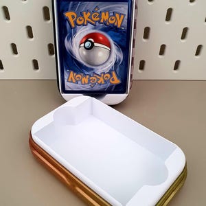 Könnte beinhalten: Eine weiße und goldene rechteckige Hülle mit weißem Innenraum, neben einer Hülle mit einer Pokémon-Sammelkarte. Die Karte zeigt das Pokémon-Logo und ein Pokéball-Design auf blauem Hintergrund. Die Hülle hat ein Schachbrettmuster.