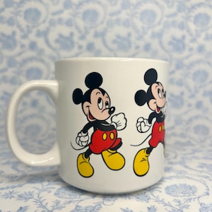 May include: White ceramic mug with a cartoon illustration of Mickey Mouse running. Mickey is shown with black ears, a red shirt, yellow shoes, and white gloves. The mug has a curved handle and is suitable for hot drinks.