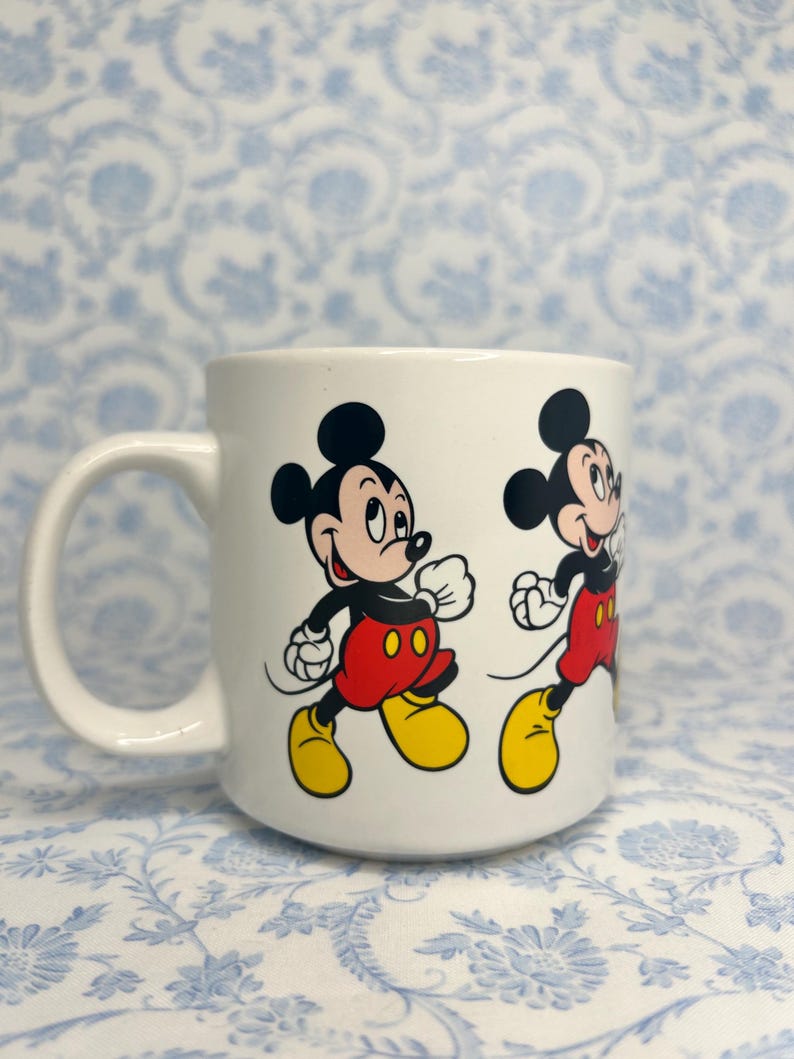 May include: White ceramic mug with a cartoon illustration of Mickey Mouse running. Mickey is shown with black ears, a red shirt, yellow shoes, and white gloves. The mug has a curved handle and is suitable for hot drinks.