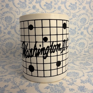 May include: A white ceramic mug featuring a black grid pattern overlaid with scattered black dots. The text Washington D.C. is written in a bold, black cursive script across the center of the grid, creating a distinct graphic design.