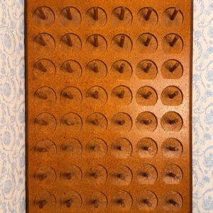 May include: A rectangular brown wooden pegboard featuring a pointed top edge and a grid of circular indentations, each containing a small vertical wooden peg. The board is constructed from a textured composite material with a uniform brown finish.