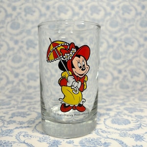 May include: A clear glass with a cartoon image of Minnie Mouse wearing a red hat and yellow dress holding an umbrella. The text "Walt Disney Productions" is printed on the bottom of the glass.