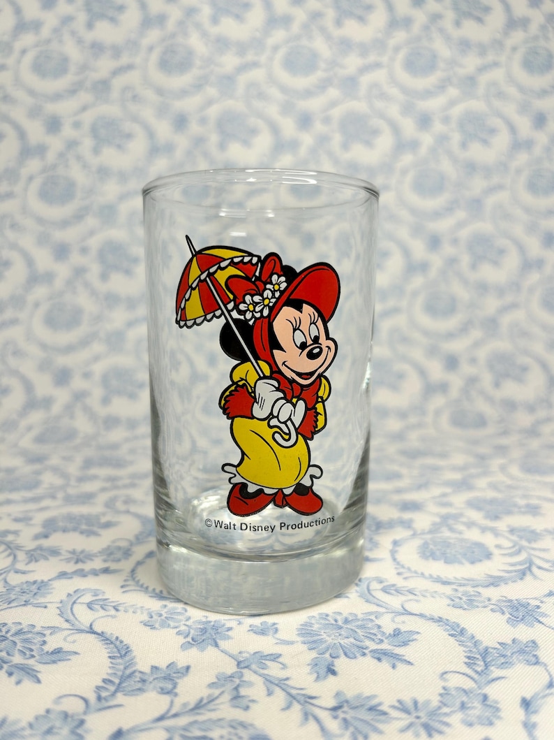May include: A clear glass with a cartoon image of Minnie Mouse wearing a red hat and yellow dress holding an umbrella. The text "Walt Disney Productions" is printed on the bottom of the glass.
