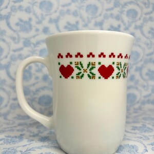 May include: White ceramic mug with a red and green pixelated design of hearts and snowflakes.