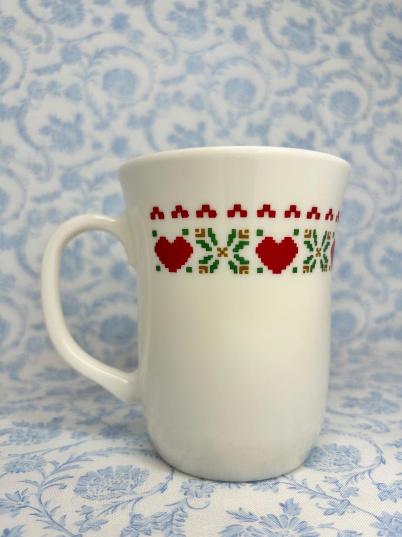 May include: White ceramic mug with a red and green pixelated design of hearts and snowflakes.
