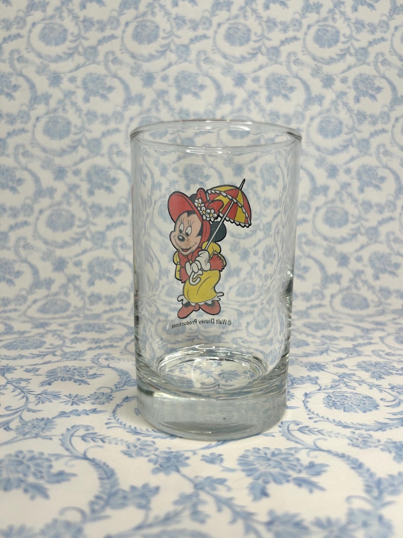 May include: A clear glass drinking glass with a cartoon image of Minnie Mouse wearing a red hat and holding an umbrella. The text "&copy; Walt Disney Productions" is printed on the glass.