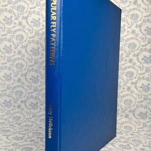May include: A blue book titled "POPULAR FLY PATTERNS" with gold lettering. The author's name, "Marry Hellekson", is also in gold. The book is set against a floral patterned background.